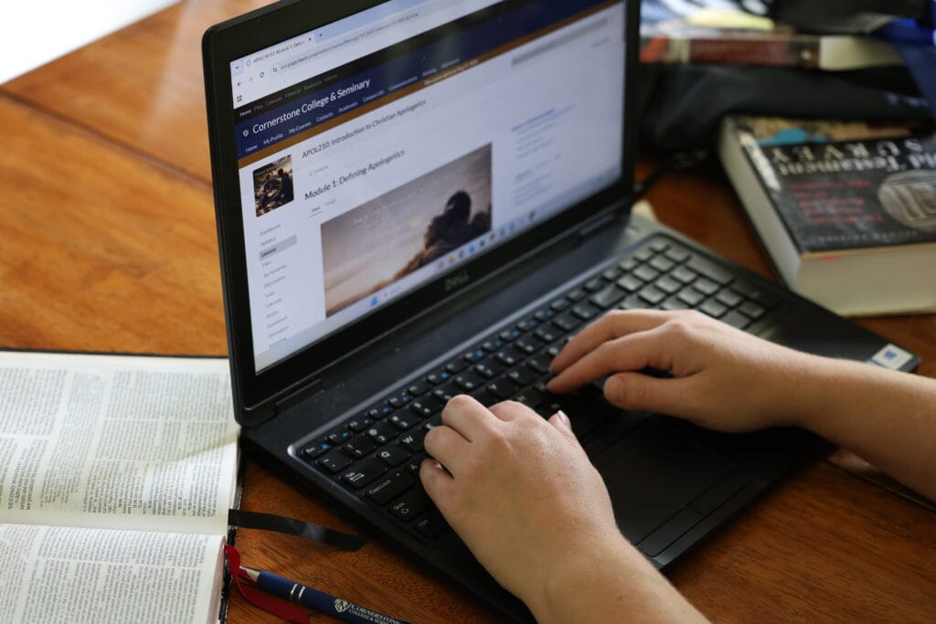 Hands typing on a laptop showing a college course page on a wood desk, with books and a pen nearby. DMIN Resources