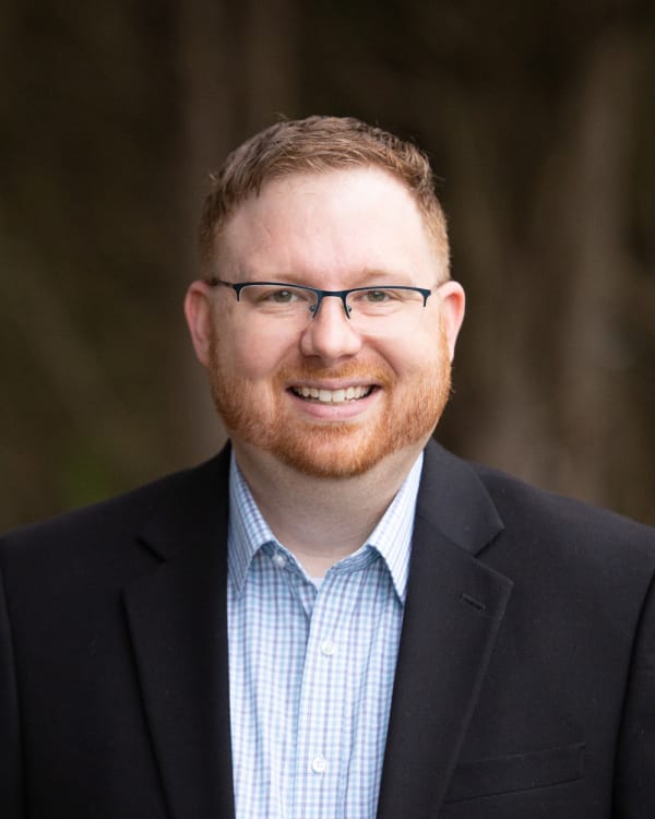 Professional headshot of a smiling man with glasses wearing a dark blazer and light blue checkered shirt, outdoors. Bryan Turner