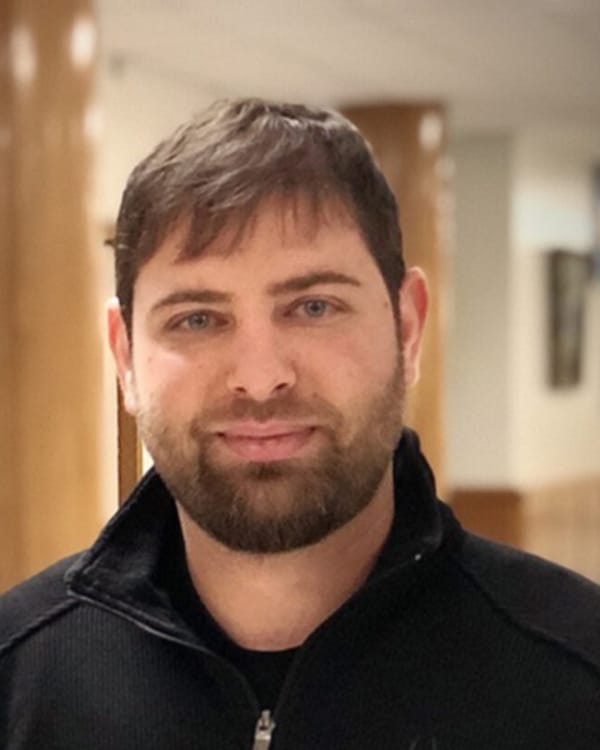 Portrait of a man with short dark hair and beard wearing a black zip sweater indoors. Bryce Woerner