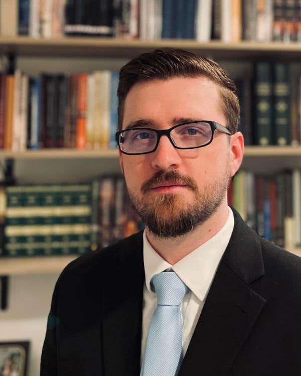Professional headshot of a man with a beard and glasses, wearing a black suit and light blue tie, in front of bookshelves in a library setting. Jason Quintern