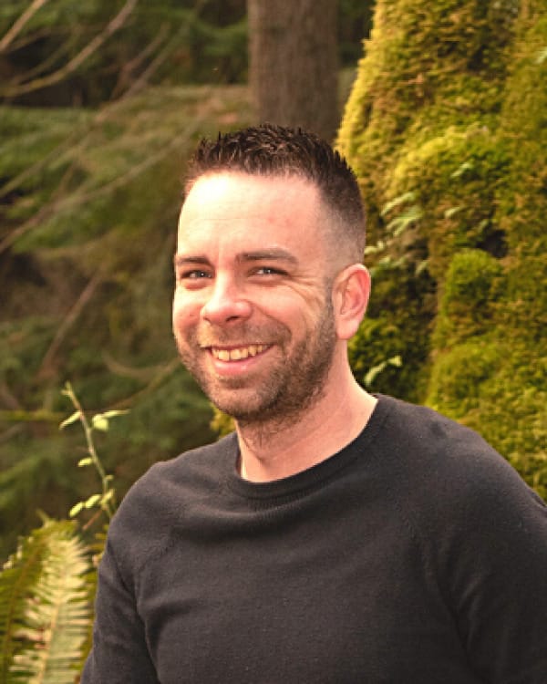Smiling man with short dark hair and stubble, wearing a black shirt, outdoors with green foliage in the background. Logan Castle