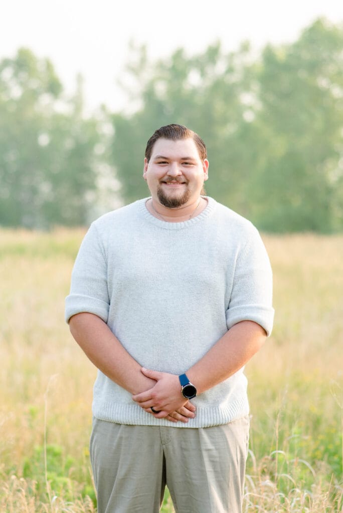 Smiling man in a light gray sweater standing in a grassy field with trees in the background. Matthew Rocke