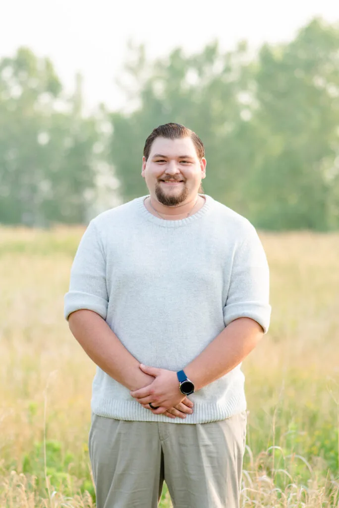 Smiling man in a light gray sweater standing in a grassy field with trees in the background. Matthew Rocke