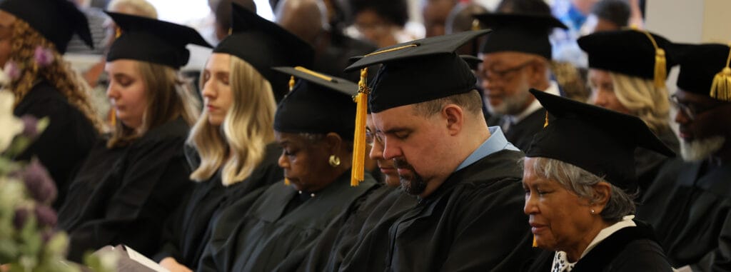 Graduates in black gowns and mortarboards seated during a graduation ceremony, some eyes closed in reflection. DMIN Resources